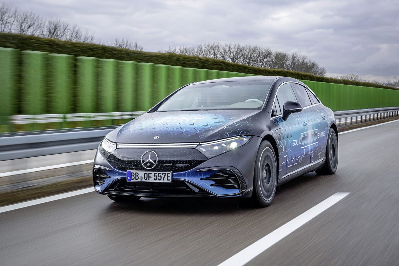 A sleek black Mercedes-Benz electric car, showcasing EV battery technology, glides smoothly along a road lined with green sound barriers under a cloudy sky.