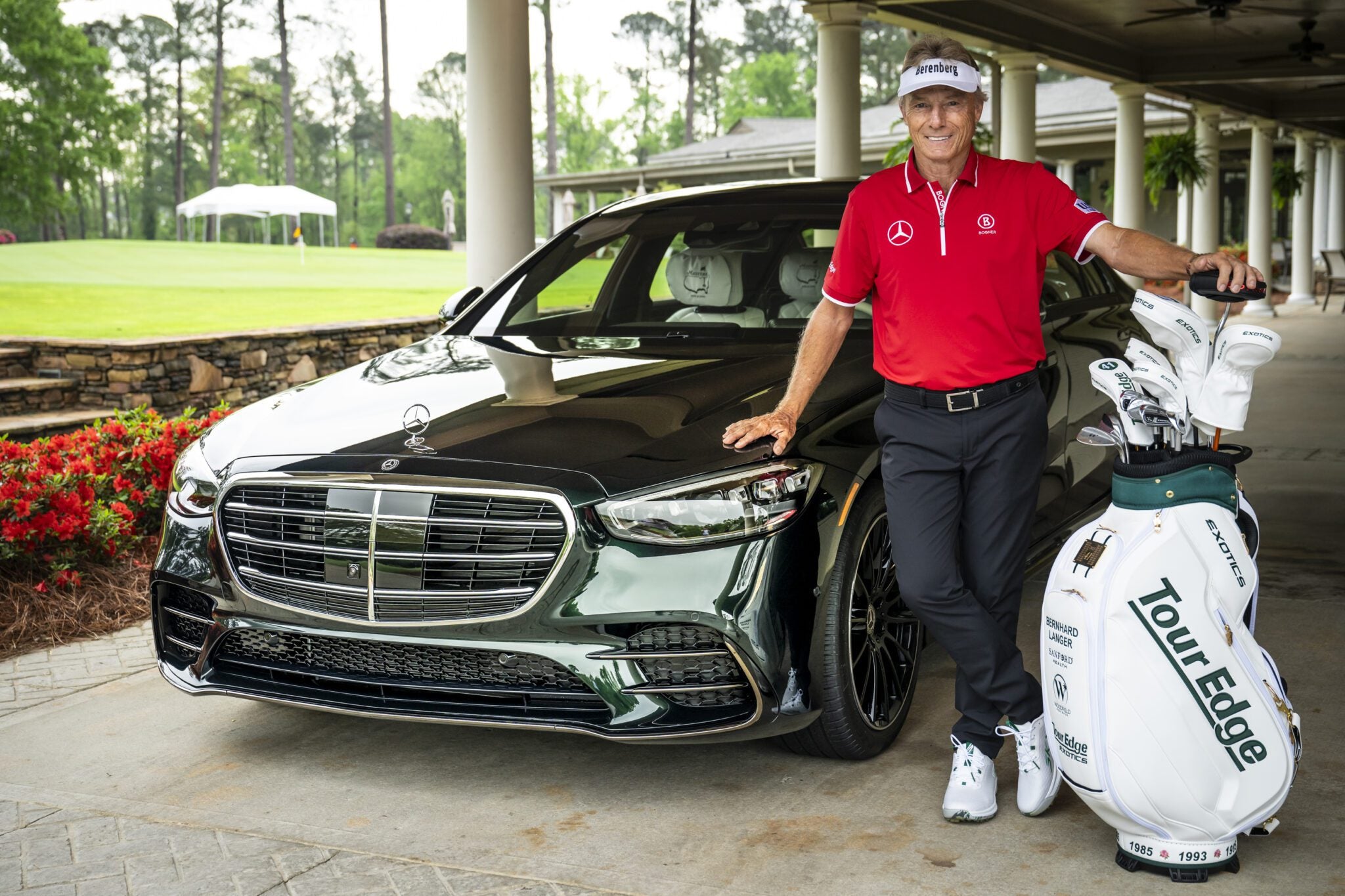 A person in a red shirt stands next to a black Mercedes-Benz sedan and a golf bag under a covered area at the prestigious Masters Augusta golf course.