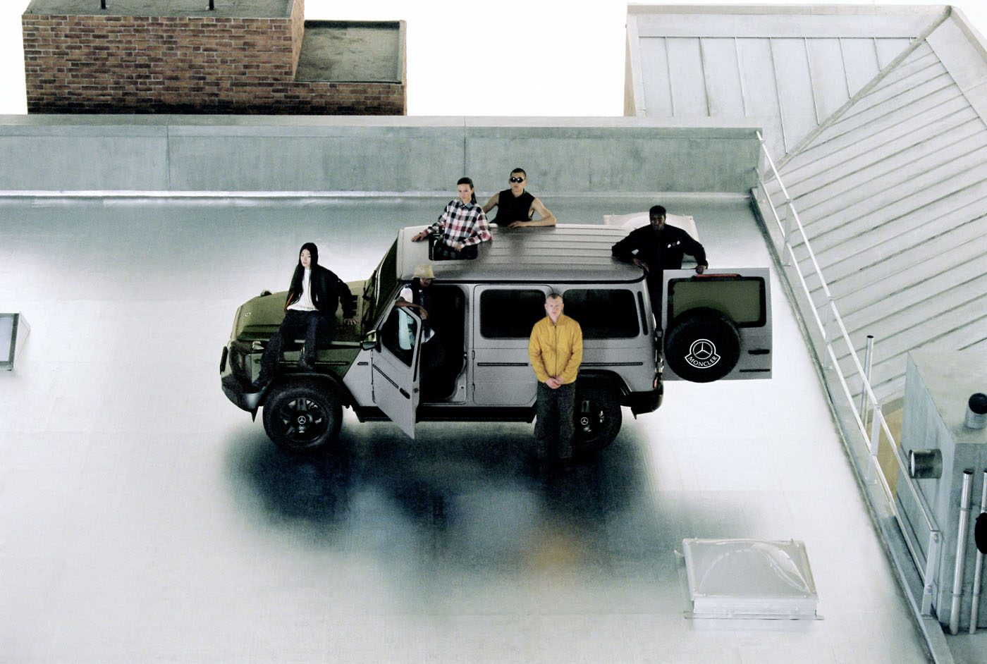 Five people gather on a rooftop with a sleek green vehicle. Some lounge atop the Mercedes-Benz, while one stands beside it. The urban backdrop of concrete and brick adds to the scene's edgy vibe, reminiscent of a stylish Moncler photoshoot.