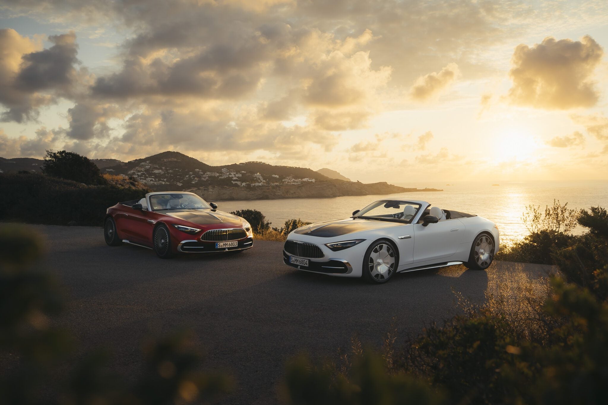 Two sporty soft-top convertibles, one red and one white, parked on a coastal road at sunset with the ocean and distant hills in the background, add a touch of Maybach luxury to the breathtaking scene.