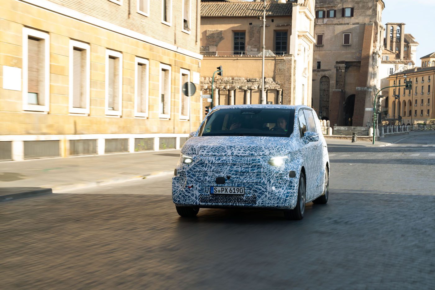 A camouflaged Mercedes-Benz prototype, possibly the VLE Electric Van, with a German license plate is being tested on a city street in Rome, with historic buildings in the background.