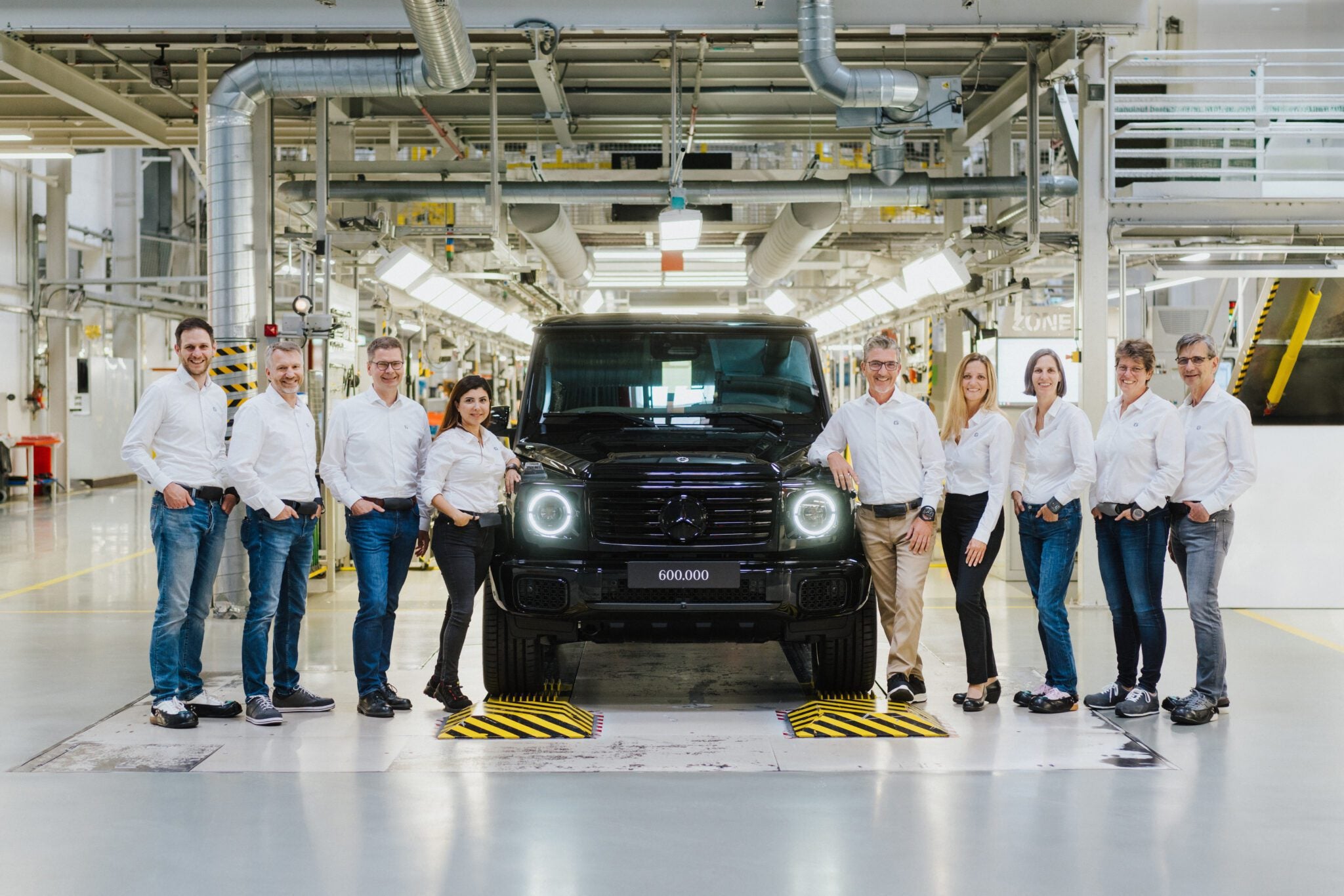 A group of ten people in white shirts and jeans stand around a black Mercedes-Benz G-Class SUV marked "600,000" inside an automotive factory.