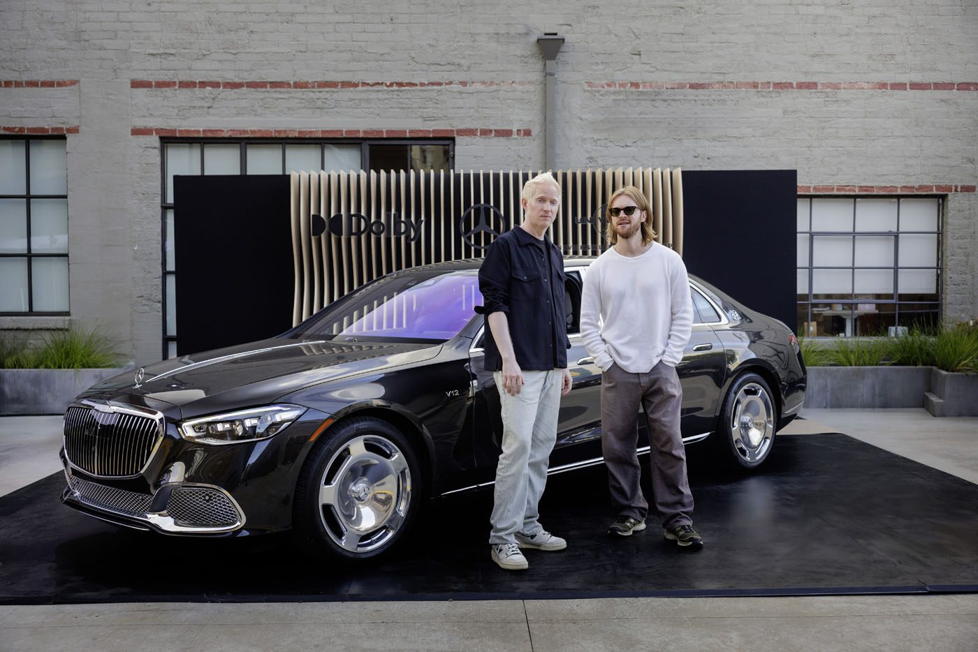 Two people stand in front of a black Mercedes-Maybach parked on a platform with a modern backdrop in an outdoor setting, resembling a rolling music studio.