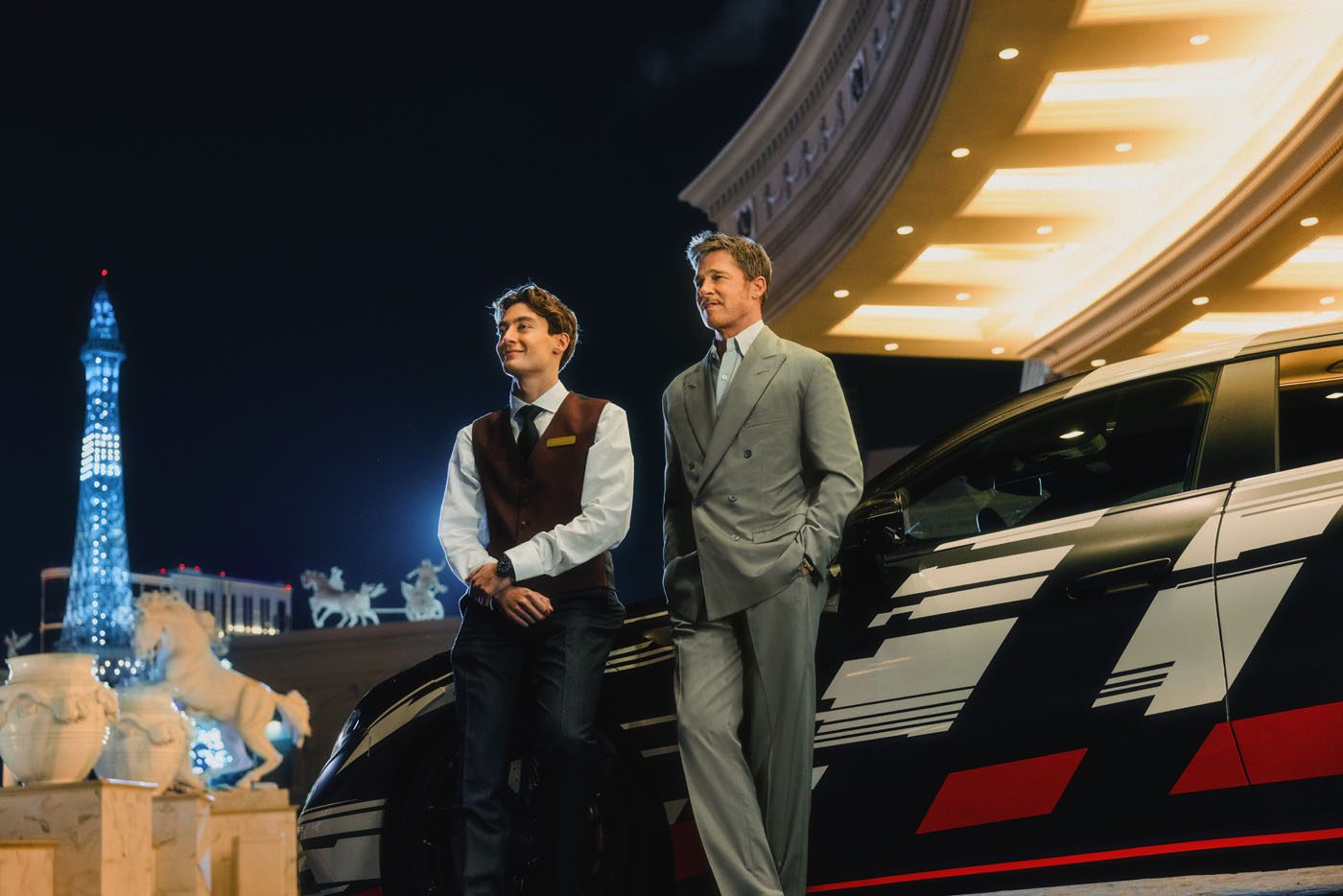 Two men in formal attire stand beside a black car at night in Vegas, with bright lights and a replica of the Eiffel Tower visible in the background.