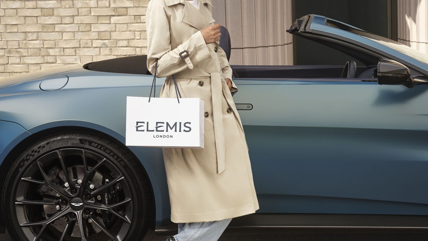 Person in a beige trench coat stands beside a blue convertible car, holding an Elemis London shopping bag.