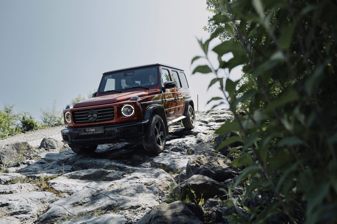A red Mercedes-Benz G-Class SUV is parked on rocky, uneven terrain with greenery and clear sky in the background, capturing the spirit of adventure that drives demand for the G-Class Experience Program.