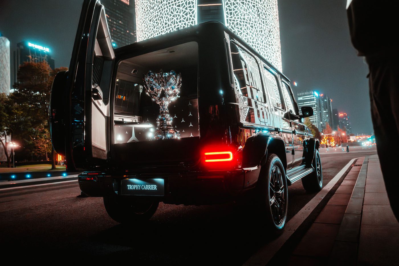 A luxury SUV with open rear doors displays a large reflective trophy in the trunk, parked on a city street at night with illuminated buildings in the background.