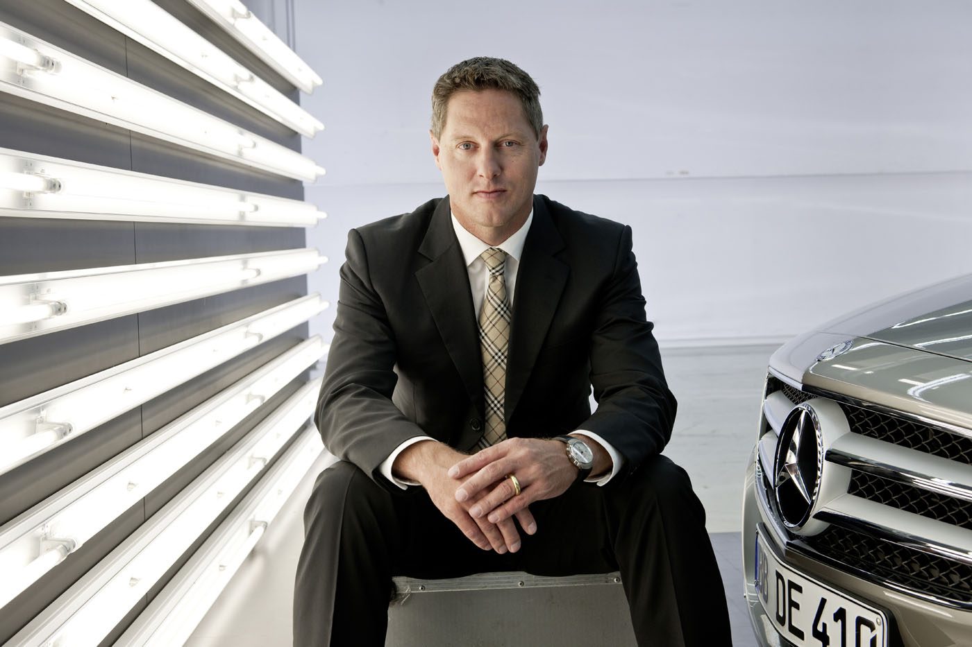 Gorden Wagener, Chief Design Officer of Mercedes-Benz, sits on a metal box beside a Mercedes-Benz car, with vertical fluorescent lights glowing on the wall behind him.