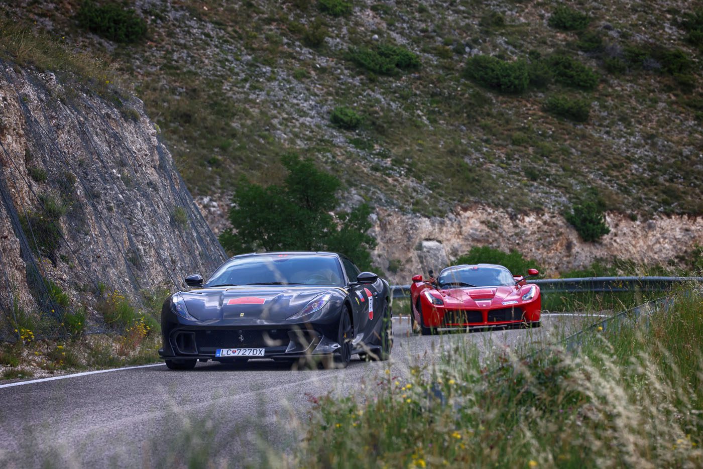 Two sports cars, a sleek black and a striking red Ferrari, race along a winding mountain road in Andalusia, bordered by rocky cliffs and lush greenery—the perfect scene for the exhilarating Cavalcade Siviglia.