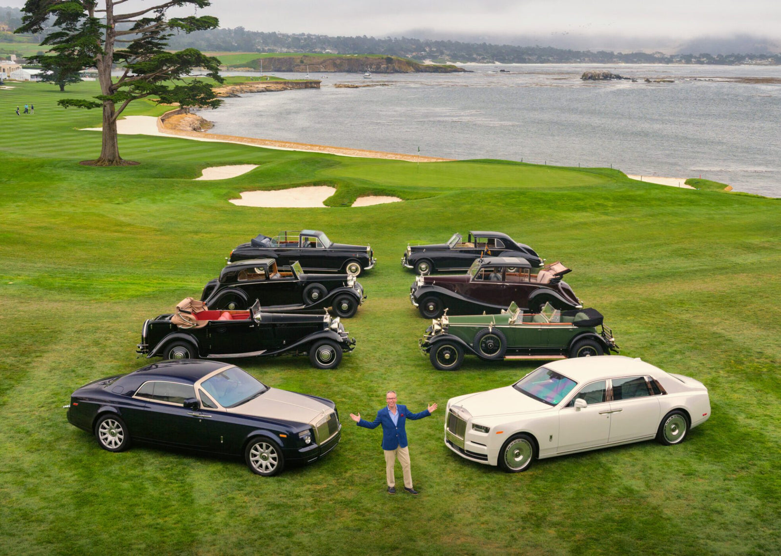 A man stands on a golf course at Pebble Beach in front of vintage and modern Rolls-Royce Phantom cars, celebrating 100 Years of automotive excellence against a stunning coastal landscape.