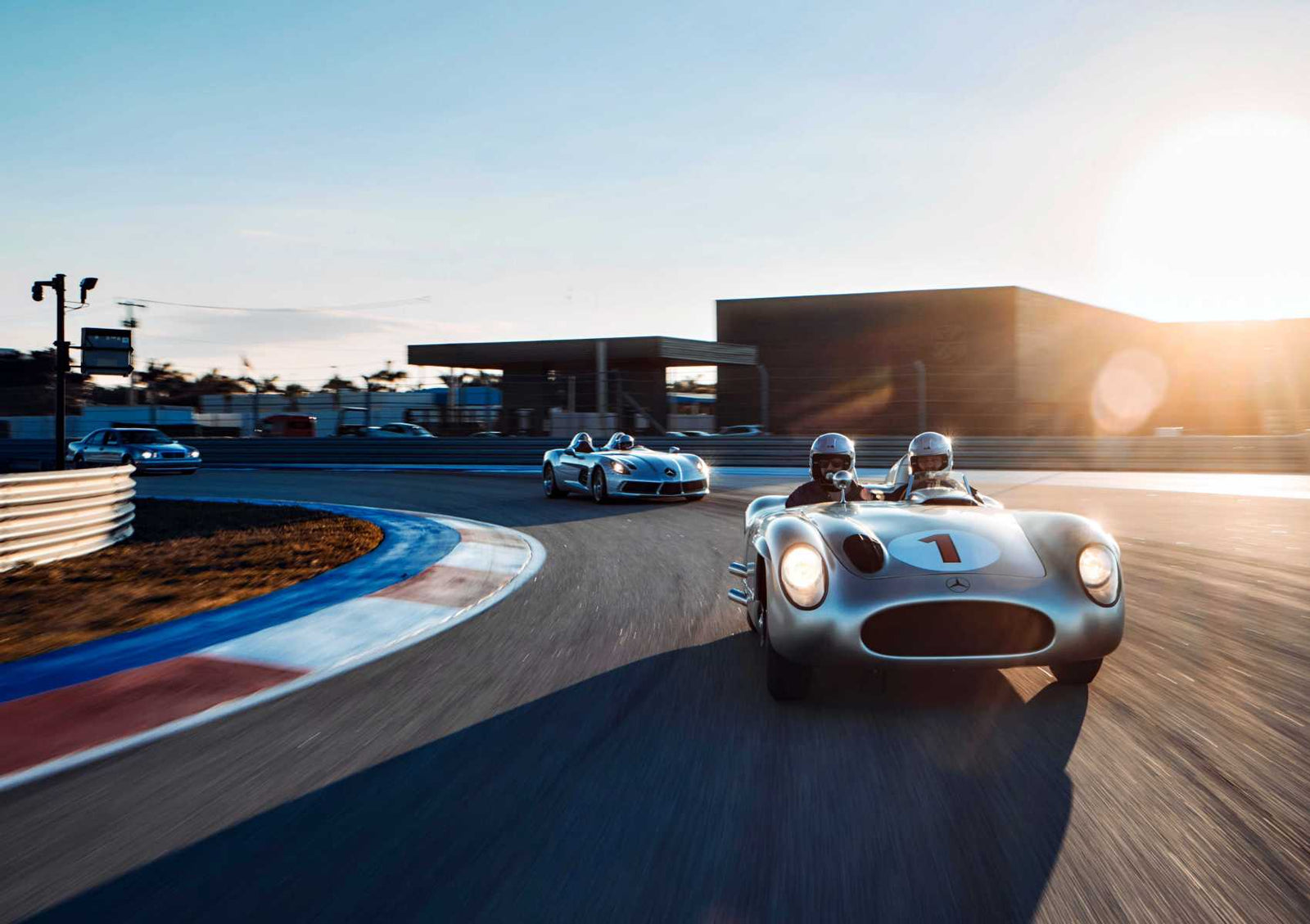 Three race cars drive on a racetrack at sunset, led by the number 1 car. Two drivers wear helmets as a modern building and barriers line the background—capturing the spirit of Must-See Mercedes-Benz Moments at Pebble Beach 2025.