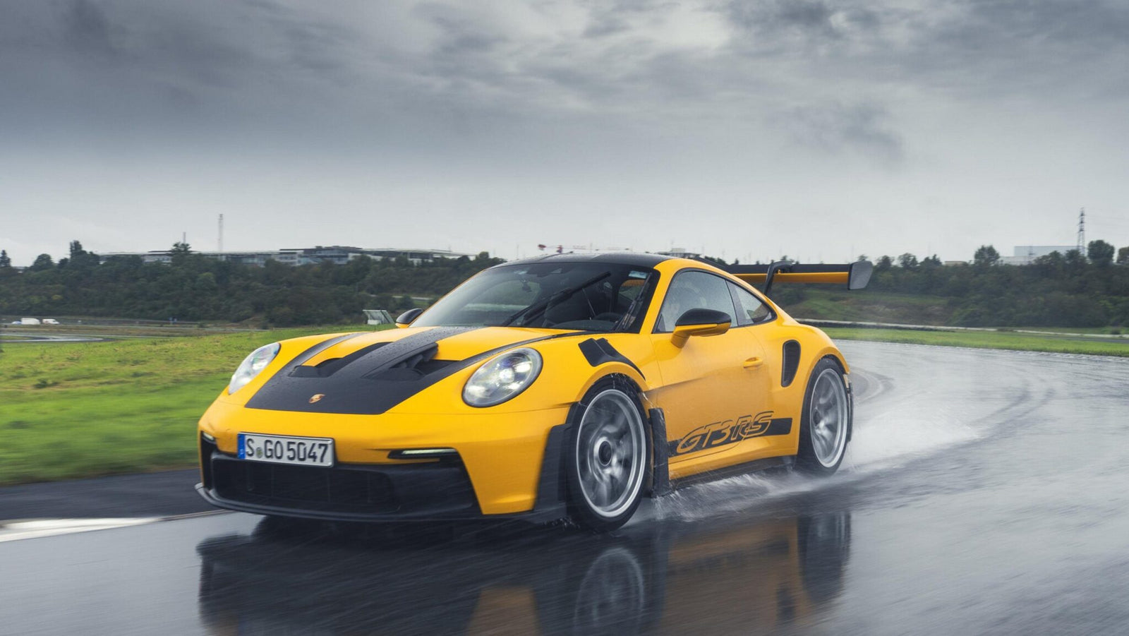 A yellow Porsche 992 911 GT3 RS zips along a wet road, its Michelin tires gripping the slick surface, while trees and buildings blur in the background.