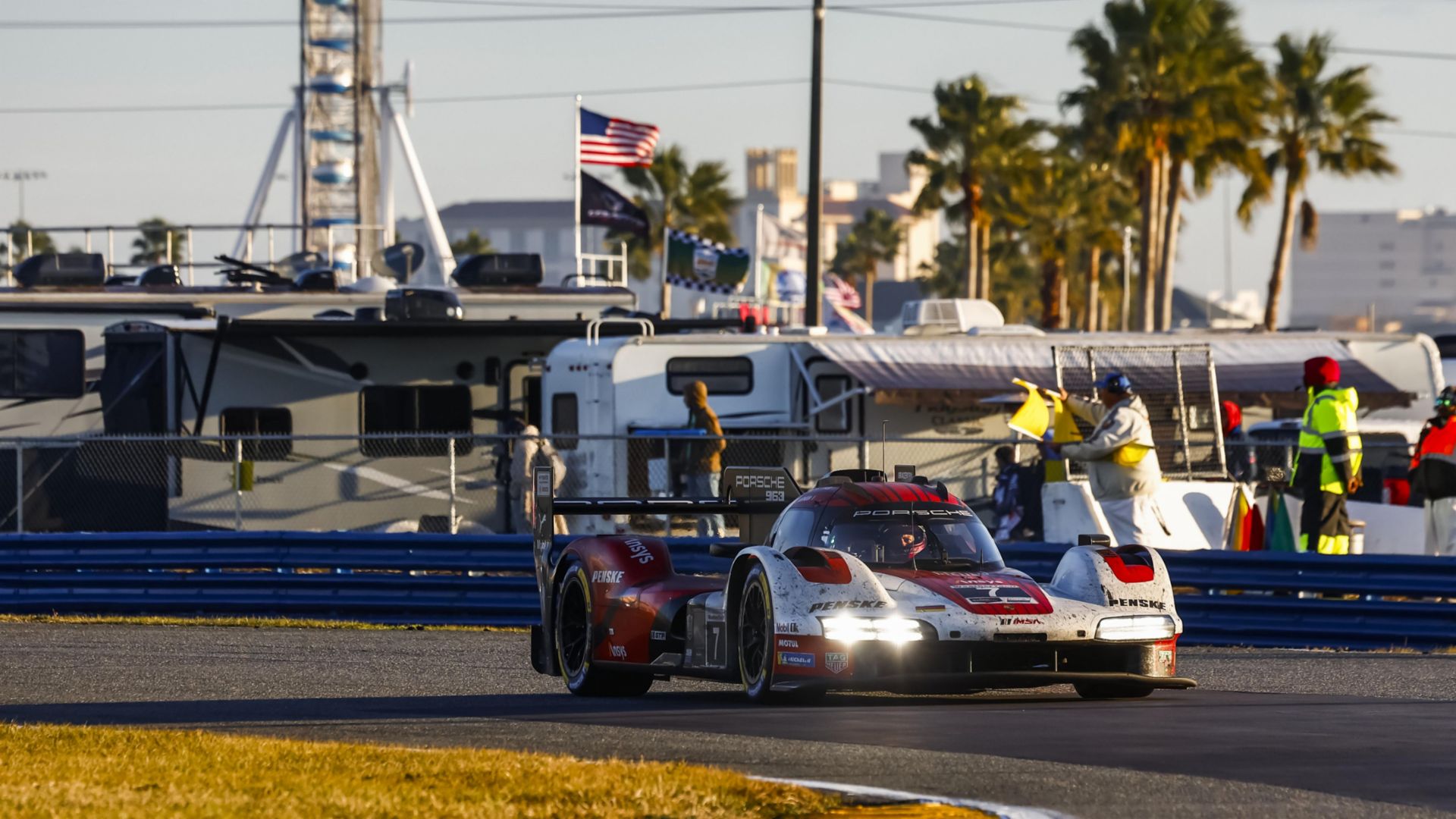 A Porsche race car speeds on the track during the 24 Hours of Daytona, with RVs, spectators, and palm trees in the background on a sunny day.