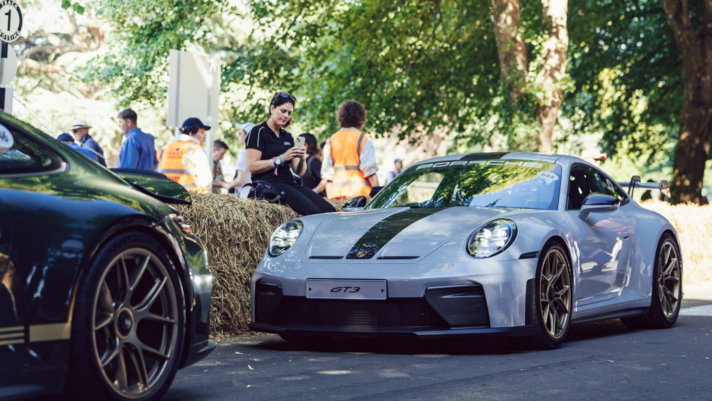 A silver Porsche 911 GT3 is parked on a shaded road at Goodwood near hay bales, with several people in the background wearing safety vests and casual clothing.