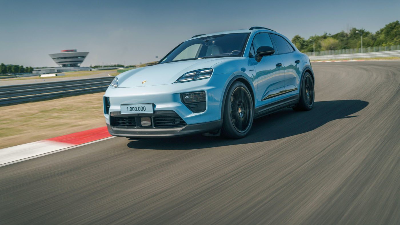 A blue Porsche Macan SUV with “1,000,000” on the license plate speeds along a racetrack, celebrating the millionth Macan, with blurred motion and a building visible in the background.