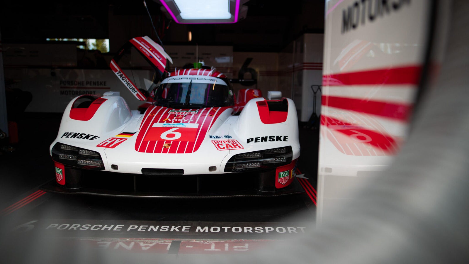 A Porsche Penske Motorsport race car with red and white livery is parked in a garage, with the driver's door open and team branding visible.