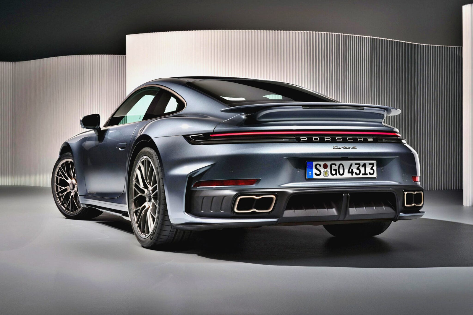 A grey Porsche 911 Turbo S, the Most Powerful Production 911: 2026 Porsche 911 Turbo S T-Hybrid Packs 701HP, is shown from the rear at an indoor display, highlighting its taillights, spoiler, rear bumper, and quad exhaust pipes.