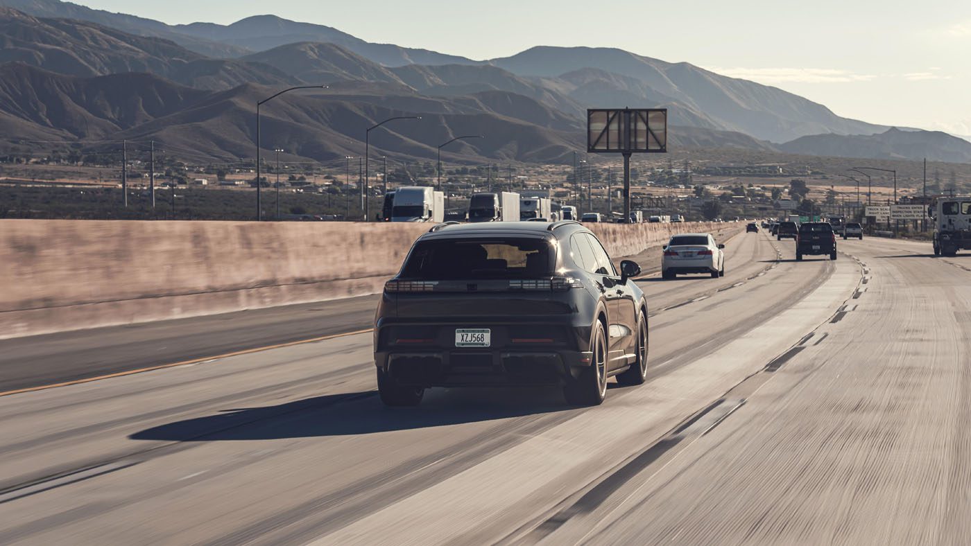 A black Porsche Cayenne Electric drives on a multi-lane highway with mountains in the background and other vehicles visible ahead under a clear sky, showcasing the future of EVs.