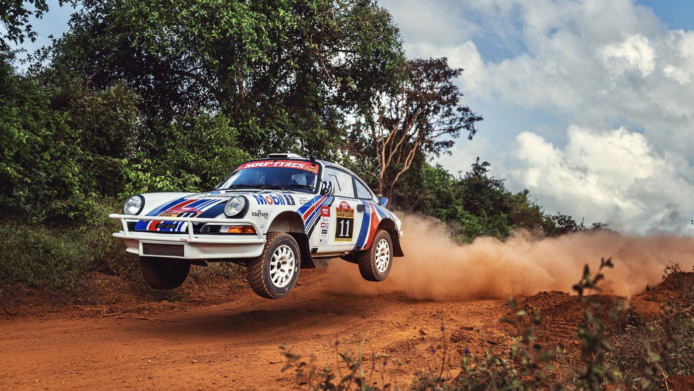 A white Porsche 911 with number 11 on the side speeds down a dirt road in the East African Safari Classic Rally, kicking up dust and lifting off the ground, surrounded by greenery and a cloudy sky.
