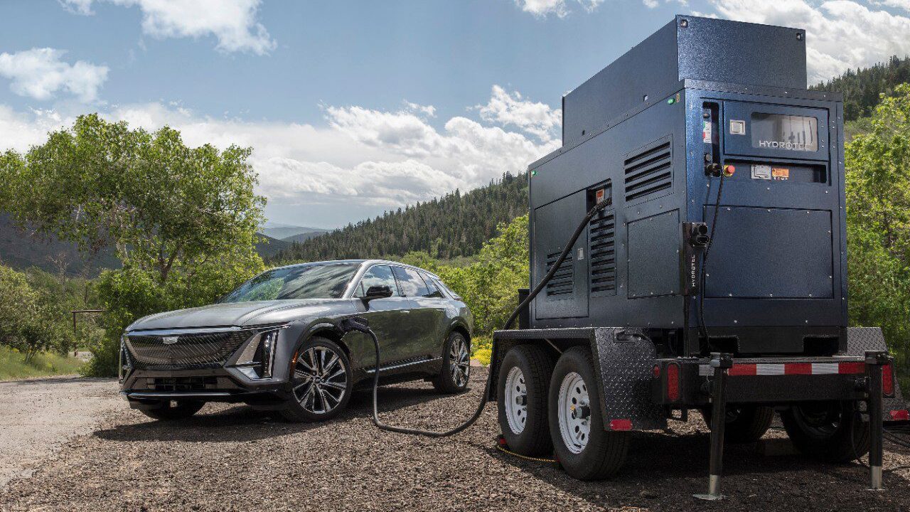 Electric car charging from a portable generator on a dirt road with trees and hills in the background.