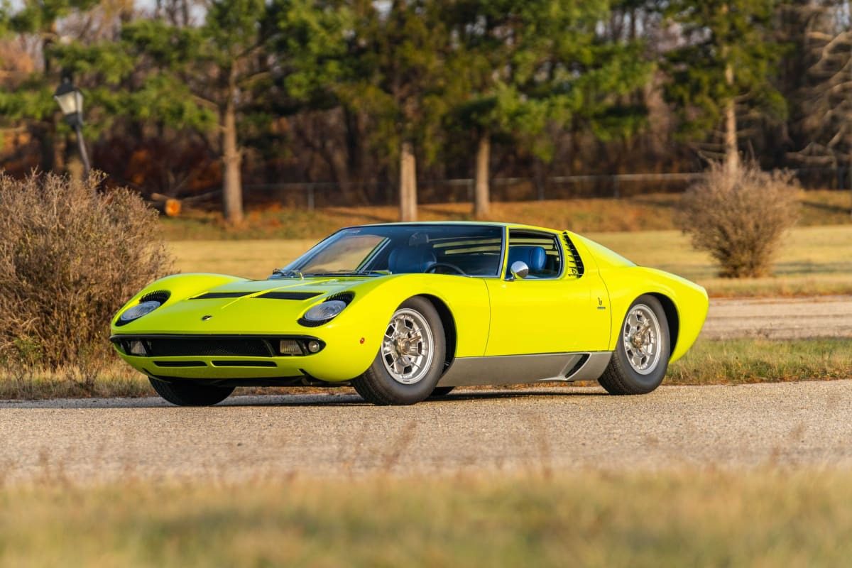 A bright yellow Lamborghini Miura sports car, a true million-dollar car, is parked on a paved road with grass, bushes, and trees in the background—ready to turn heads at the Monterey 2025 Auction by Mecum.