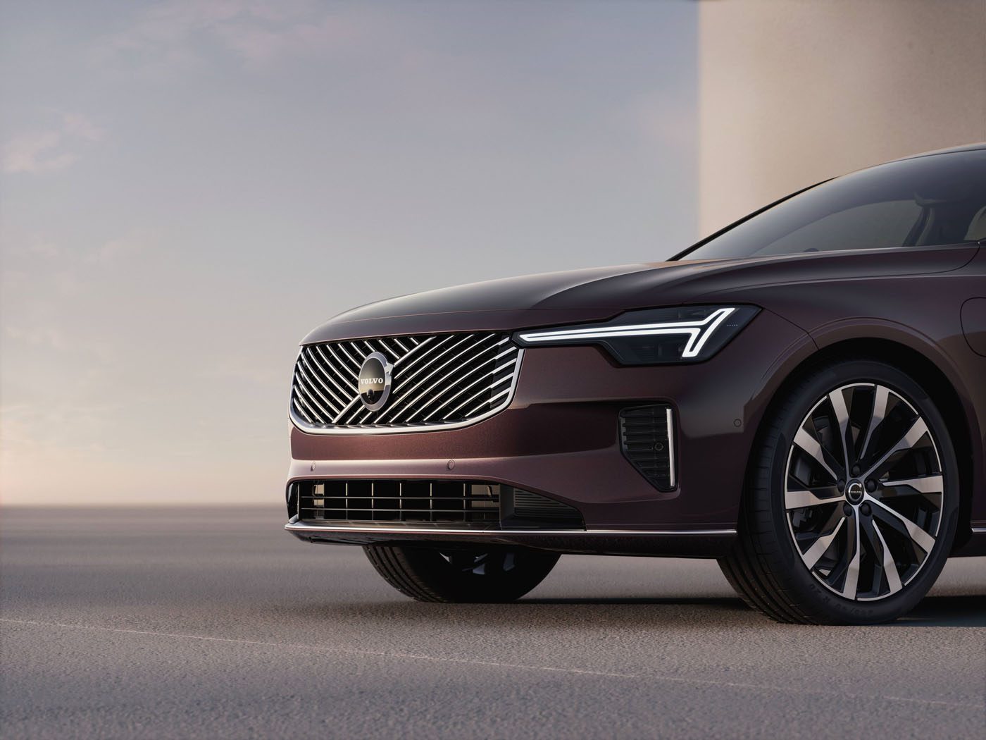 Close-up of the front side of a modern maroon Volvo Hybrid S90, showing the grille, headlights, and part of a wheel on a paved surface.