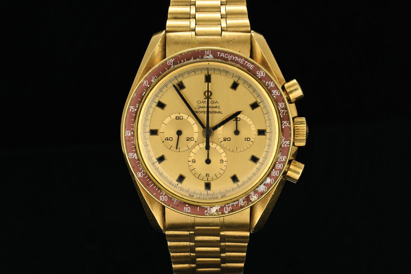 The dazzling 18k Gold Omega Speedmaster watch, reminiscent of the timepiece Neil Armstrong wore, boasts a tachymeter scale on a striking red bezel, three sub-dials, and a link bracelet. Its elegance is perfectly captured against a deep black background.
