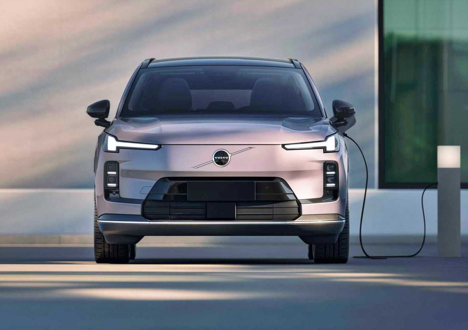 Front view of a light pink Volvo electric SUV parked and charging at a station next to a modern building, reflecting Volvo’s global plug-in hybrid sales crossing one million.