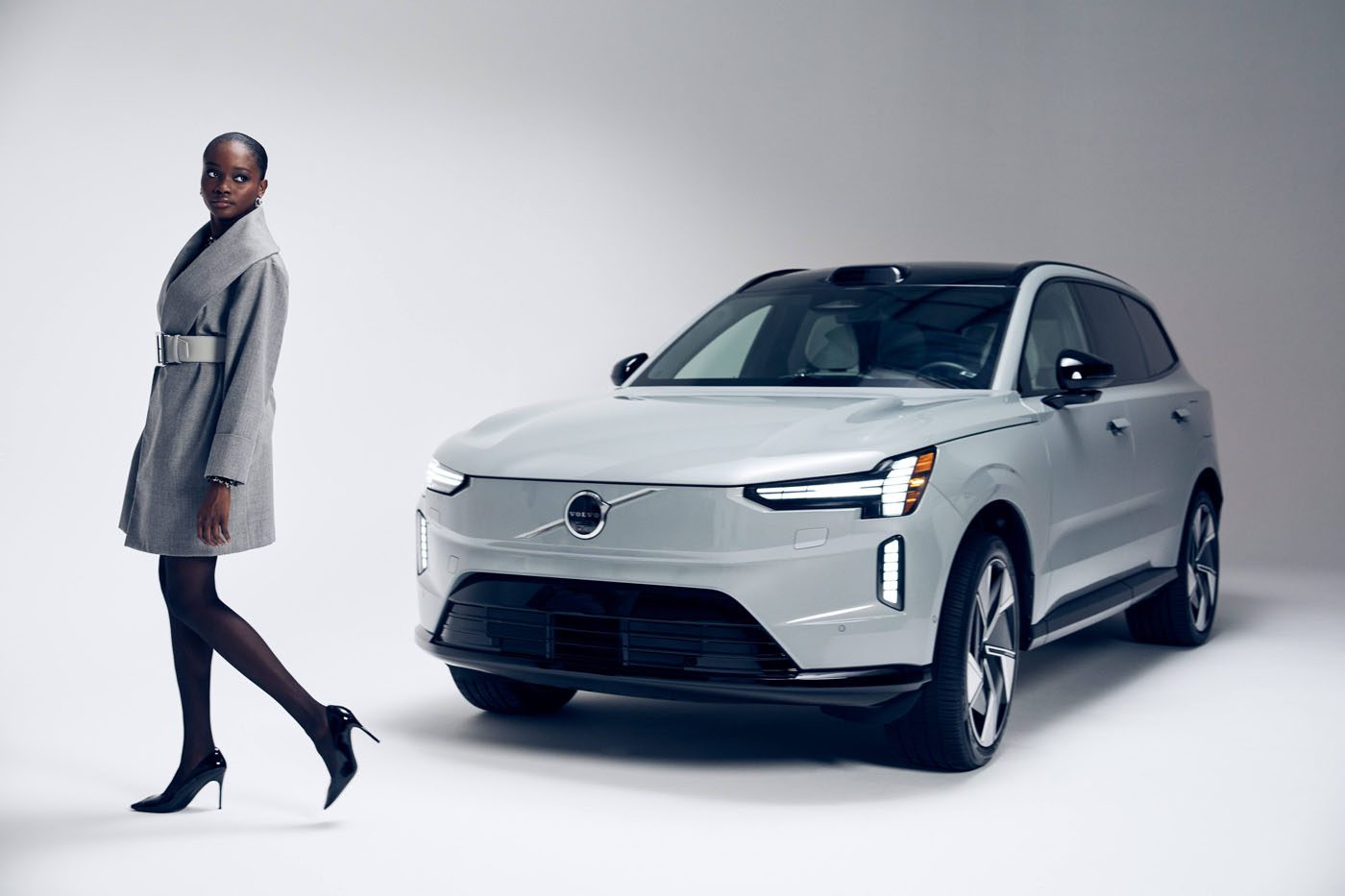 A woman in a Sergio Hudson grey coat and heels stands next to a silver Volvo EX90 SUV in a studio with a plain light background.