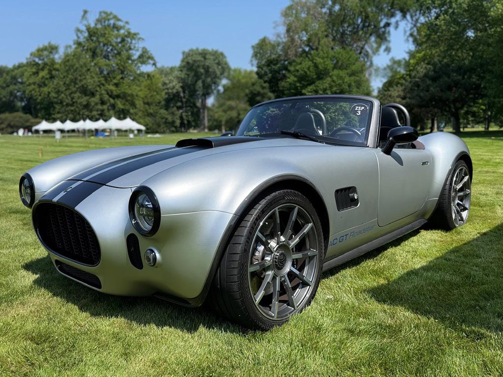 A silver convertible sports car with black stripes is parked on grass, with trees and tents in the background under a clear sky.