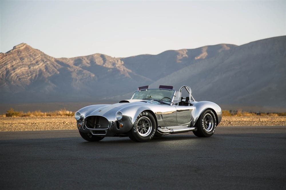 A silver Shelby 427 Cobra sports car sits elegantly parked on a road, framed by the majestic mountains in the background.