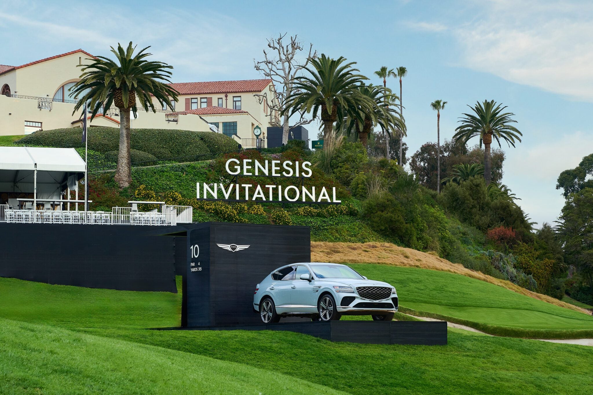 A silver car is showcased on a raised platform near a golf course, with "2025 PGA Invitational" elegantly scripted on the backdrop, surrounded by palm trees and buildings, symbolizing support for Wildfire Relief.