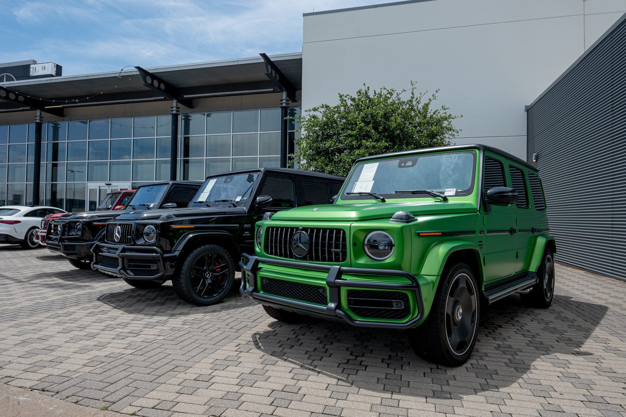 Several black and one green luxury SUVs are parked outside a modern Mercedes-Benz dealership building on a paved area.