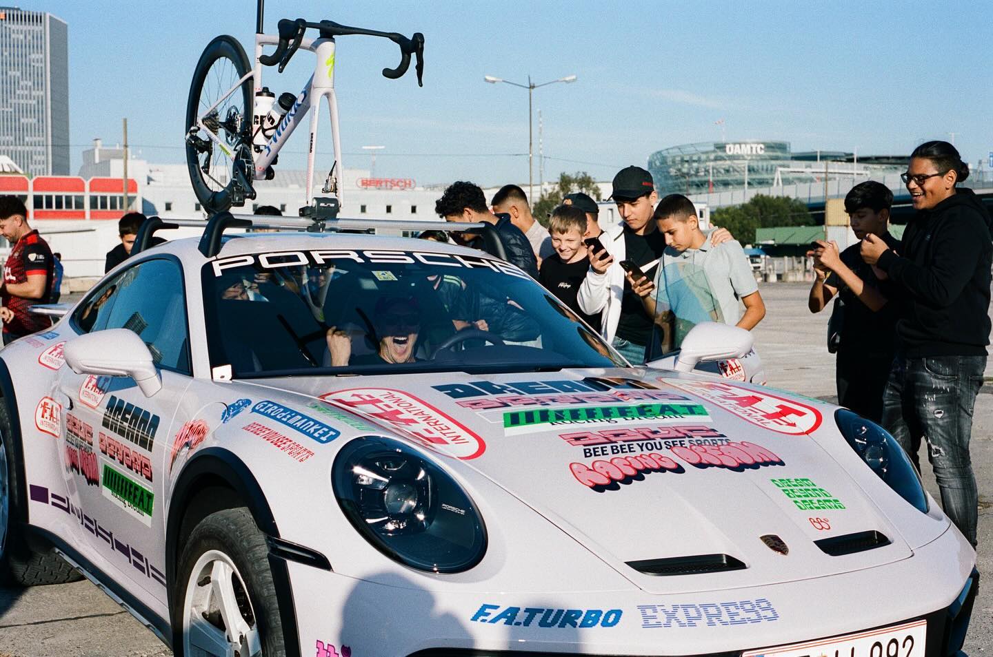 A group of people gather around an exclusive release—a Porsche 911 Dakar—adorned with various decals and a Specialized Bike on the roof, eagerly snapping photos with their phones.