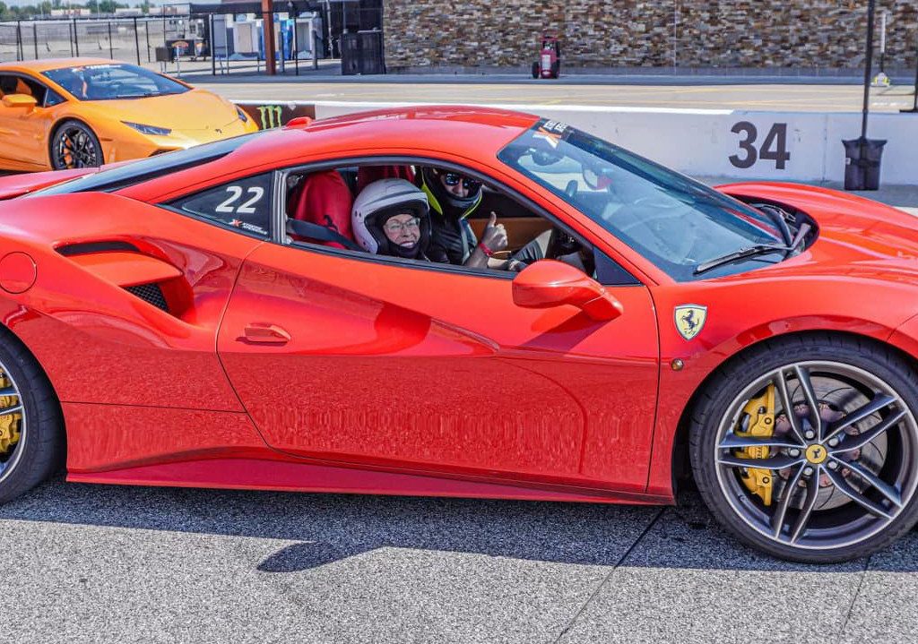 A 94-year-old sits inside a red Ferrari 488 GTB on the racetrack, clutching a phone. An orange sports car looms in the background, capturing the essence of a lifelong racing dream coming to life.