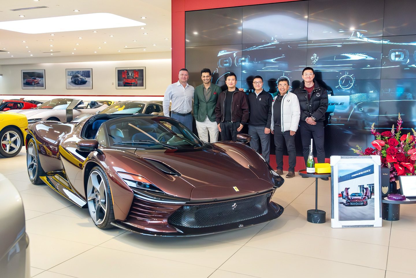 A group of six people stands behind a sleek, dark brown convertible in an upscale showroom filled with other luxury vehicles, including the impressive Ferrari Daytona SP3.