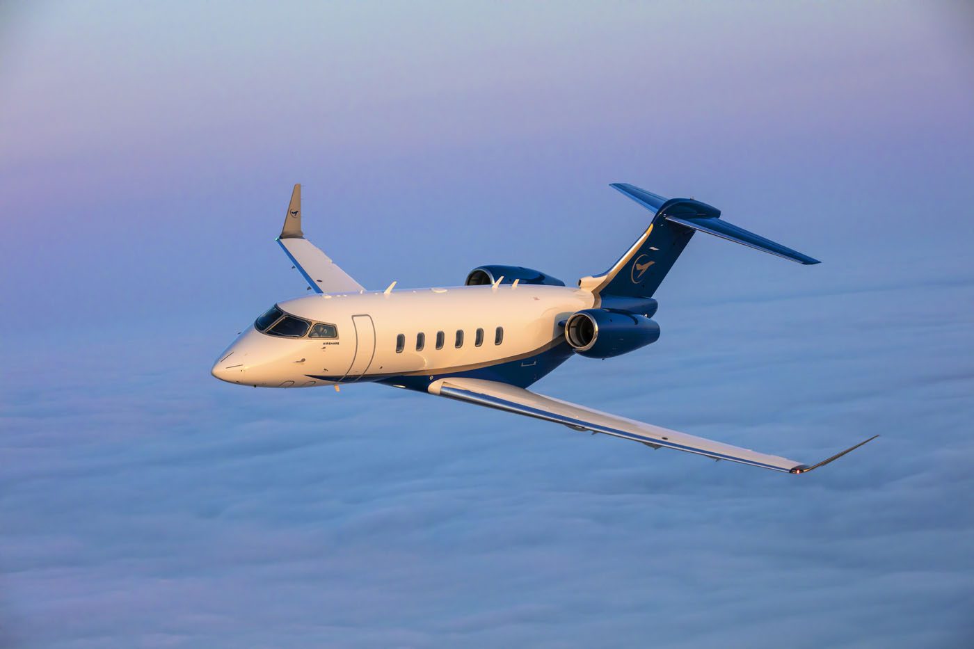 A blue and white Airshare private jet flies above a blanket of clouds during sunset or sunrise.