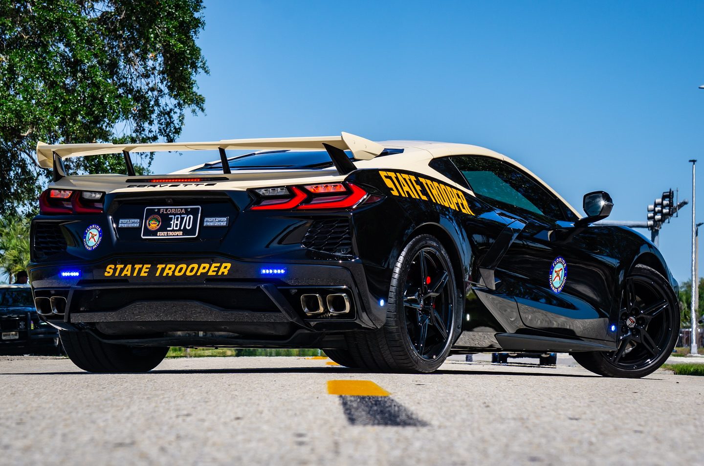 A sleek black and white C8 Corvette, adorned with a spoiler, emergency lights, and the Florida Highway Patrol insignia on its sides, is parked on a scenic road amidst trees under a clear blue sky.
