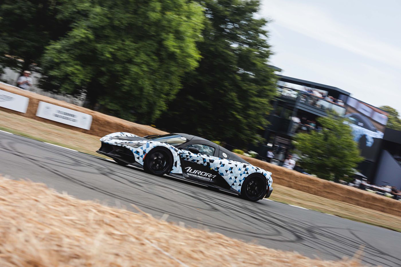 A Zenvo Aurora sports car with geometric camouflage and "Aurora" branding makes its global debut on a racetrack bordered by hay bales, with trees and a multi-level building in the background.