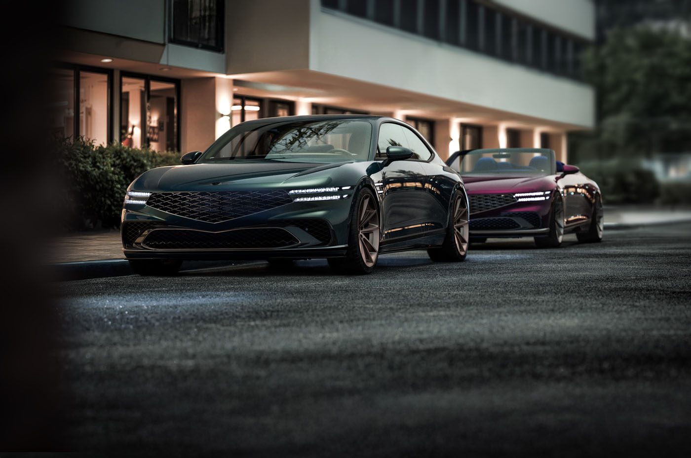 Two sleek, modern vehicles, including a Genesis X Gran Coupe with angular designs and illuminated headlights, stand parked on a city street in front of a building boasting large windows.