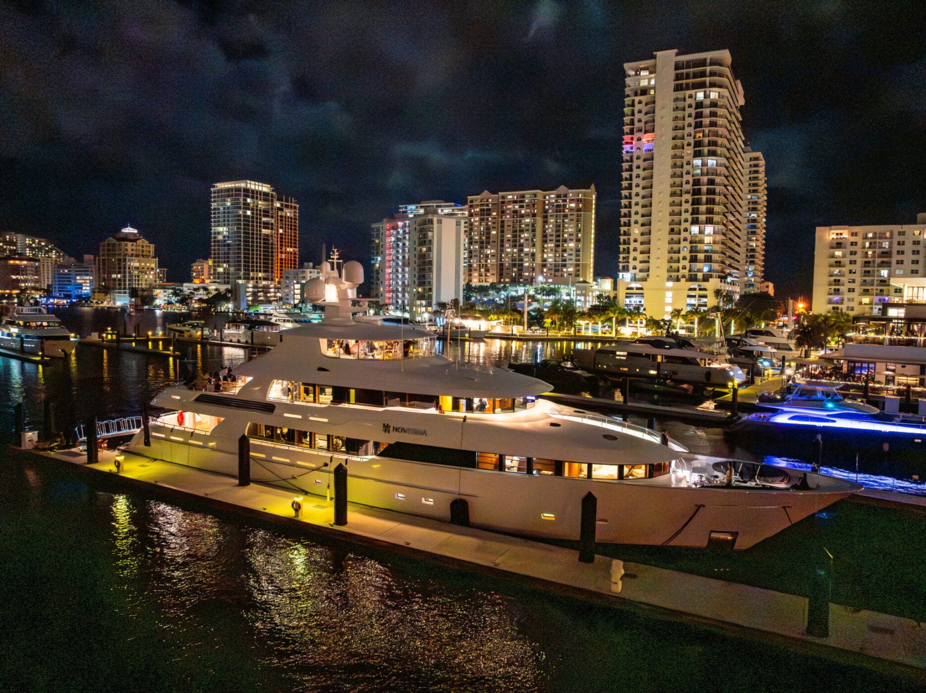 A large, illuminated Delta Marine Noverra yacht docked at a marina at night, with tall city buildings and lights reflected on the water—an iconic scene of five-star living and luxury yacht charter perfection.