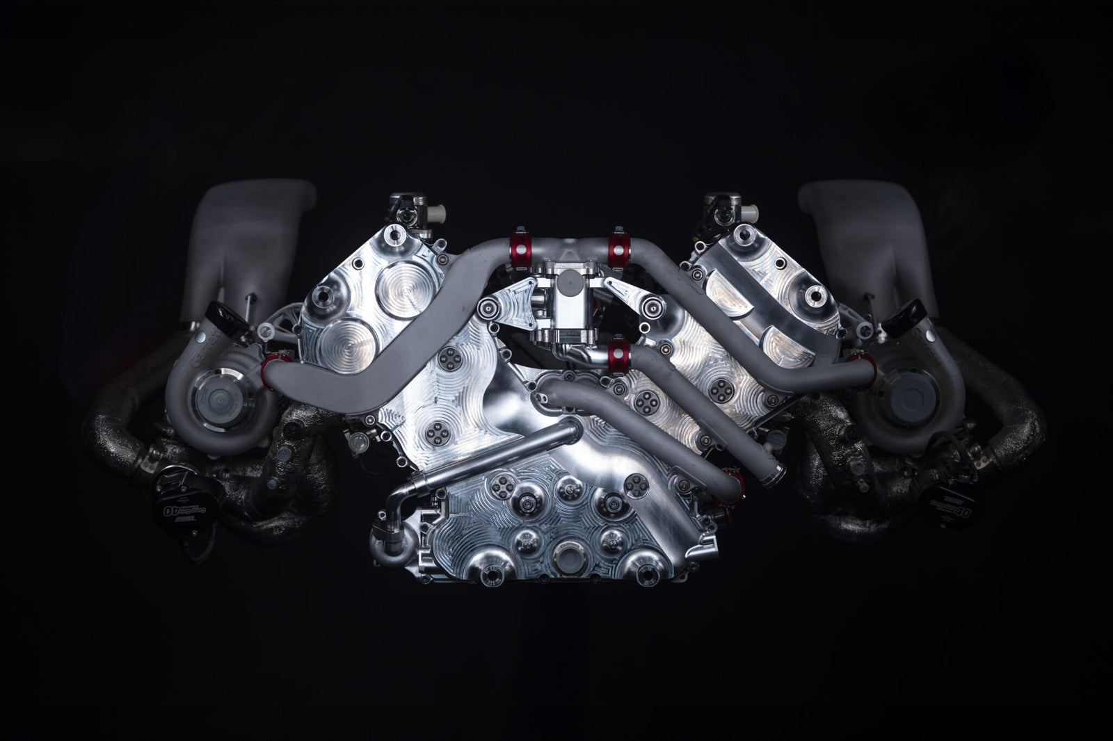 A metallic V-shaped twin-turbocharged engine is photographed against a black background, showcasing its complex pipes and components.