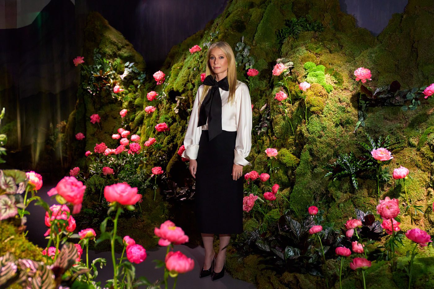 A woman stands in front of a green, mossy backdrop decorated with pink flowers at Genesis House’s The Forest Within, wearing a black skirt, white blouse, and black shoes—evoking Gwyneth Paltrow’s effortless elegance.