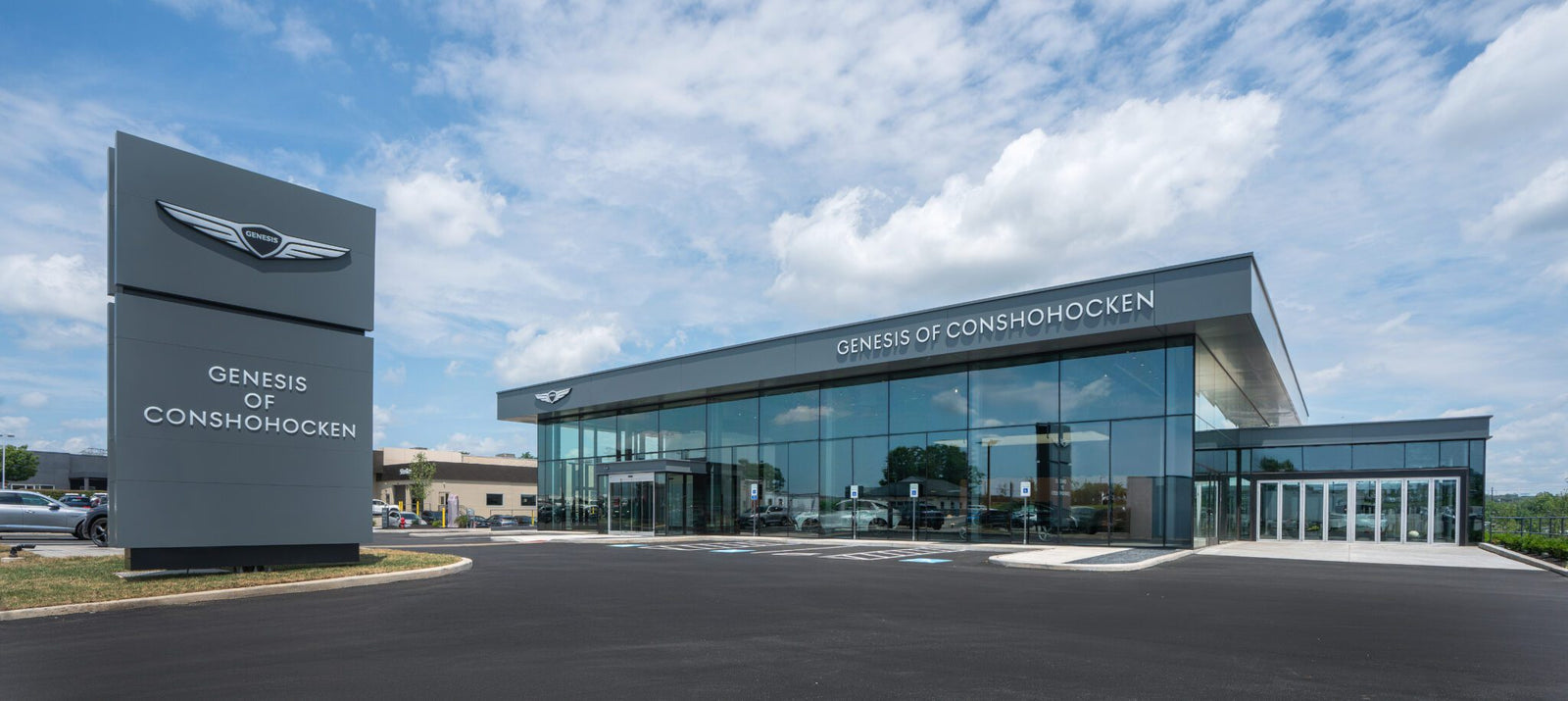 Modern car dealership building with large windows and a sign reading "Genesis of Conshohocken" under a partly cloudy sky.