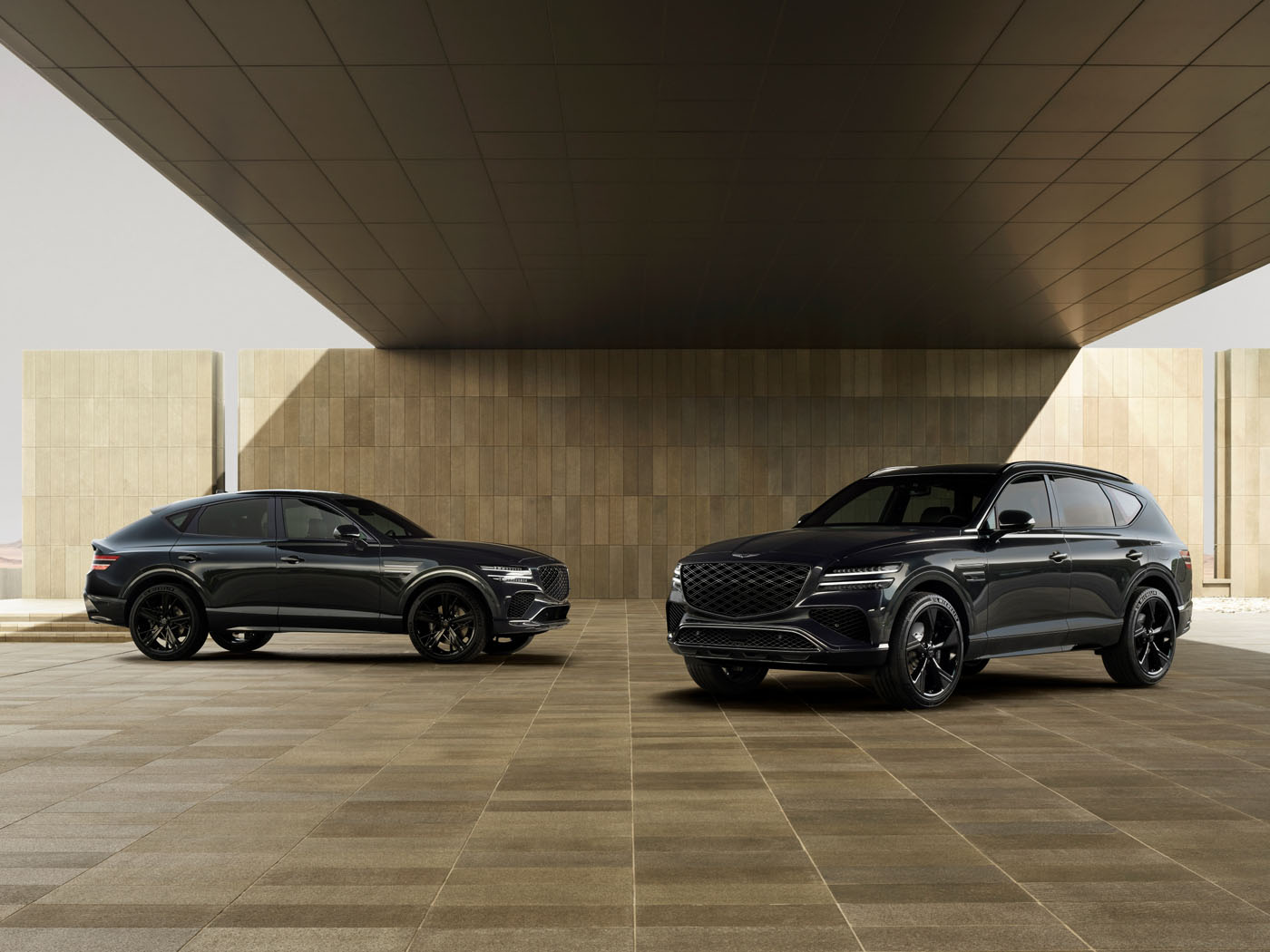 Two black SUVs are parked on a tiled surface under a large, modern overhang with beige stone walls in the background.