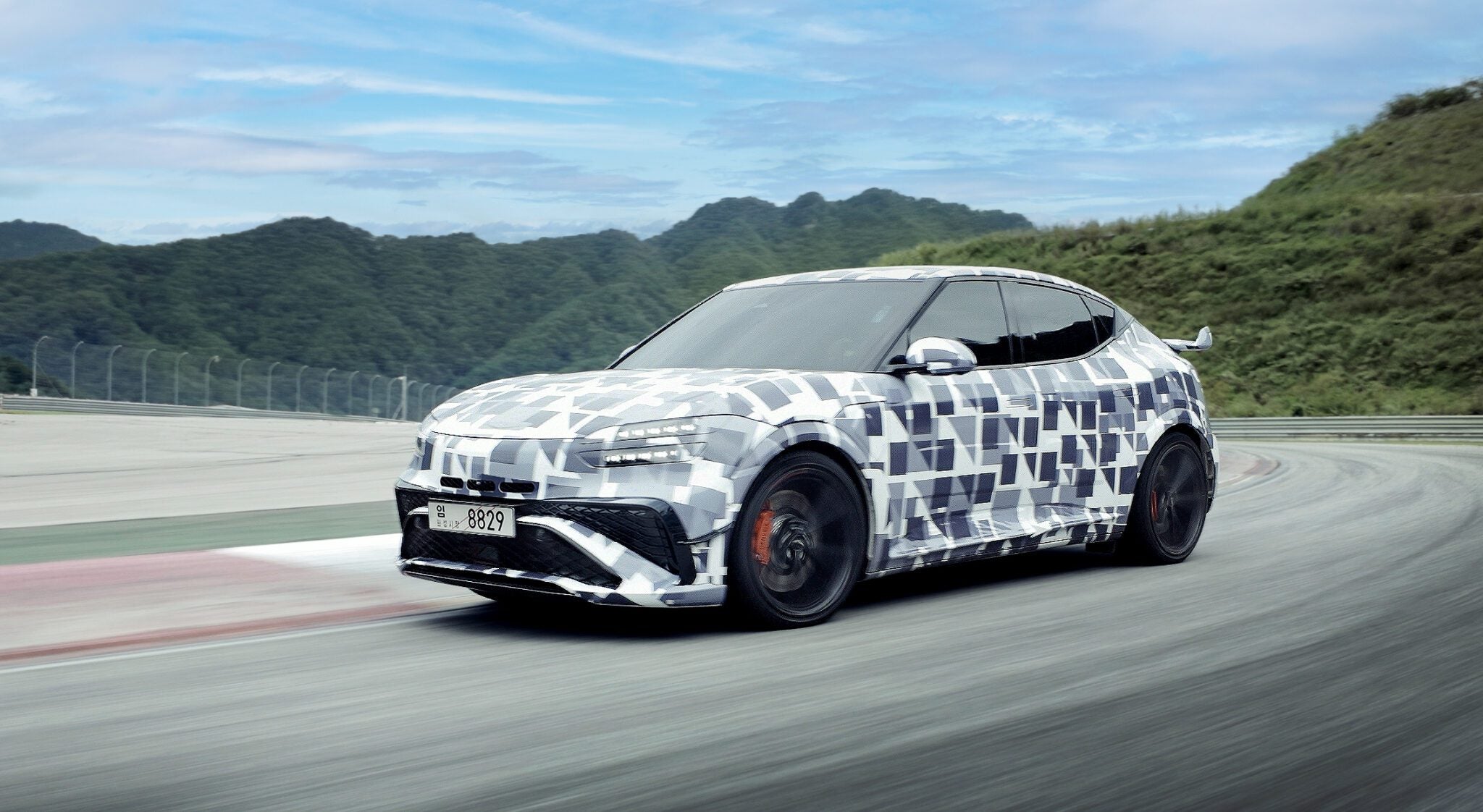 A camouflaged car with geometric wrap drives on a racetrack with green hills and a blue sky in the background.