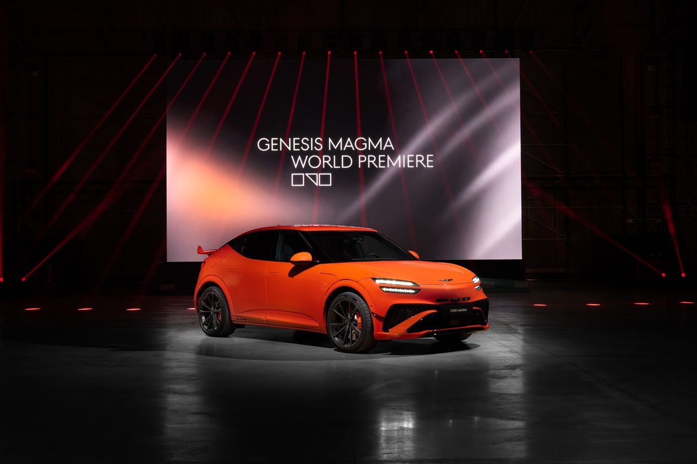 An orange Genesis GV60 Magma car is displayed in front of a large screen reading "Genesis Magma World Premiere" in a dark indoor venue with red accent lighting, showcasing the latest production model.
