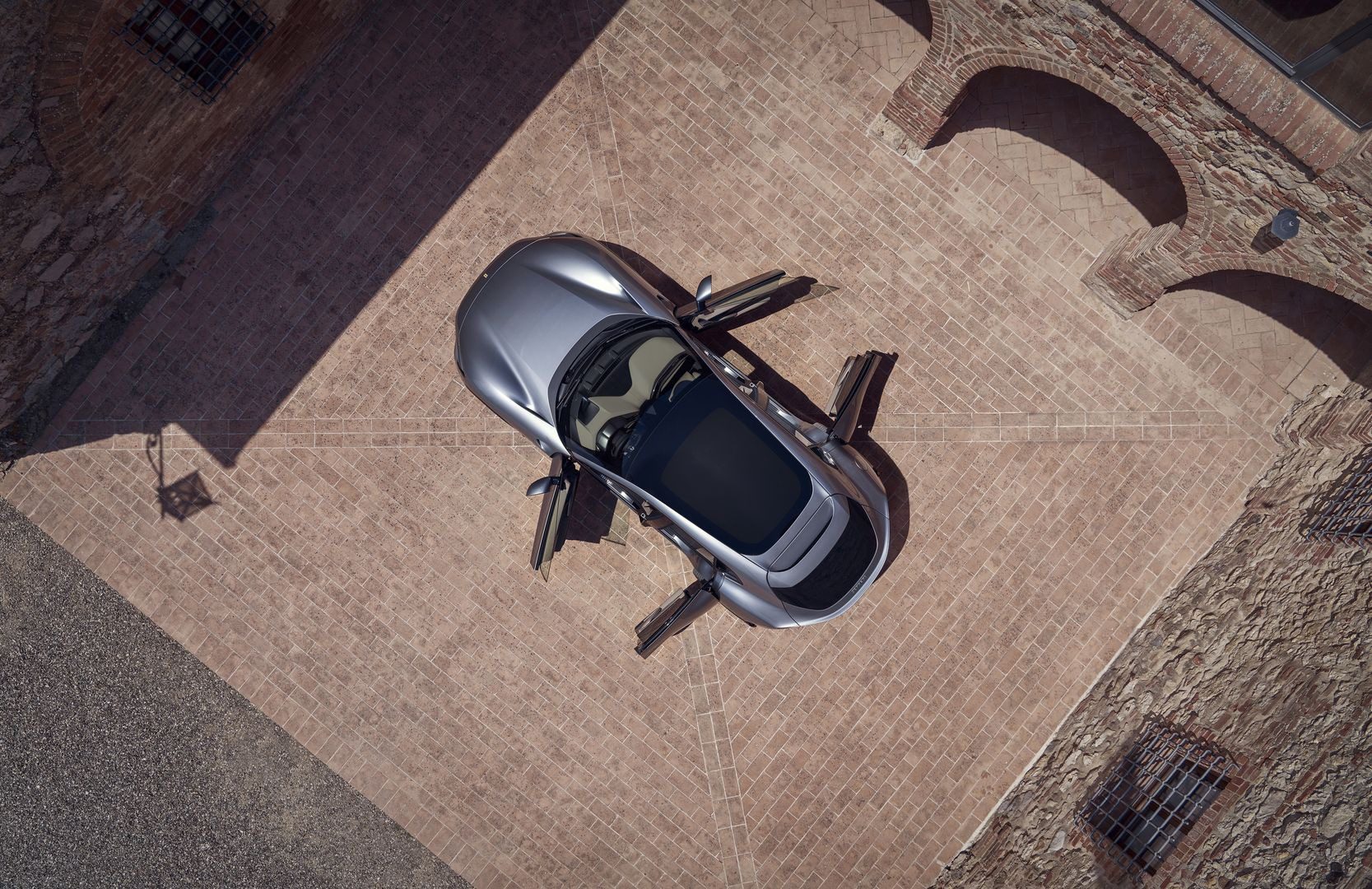 Overhead view of a sleek silver Ferrari sports car with its doors open, parked on a brick driveway surrounded by brick buildings.