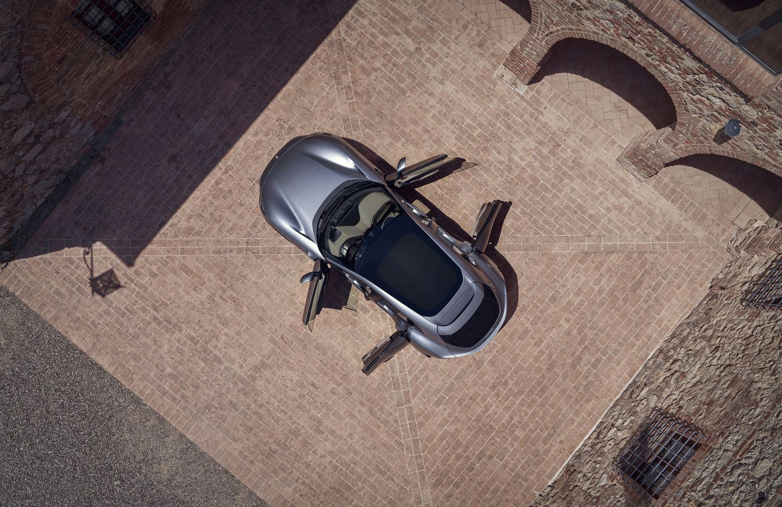 Overhead view of a sleek silver Ferrari sports car with its doors open, parked on a brick driveway surrounded by brick buildings.