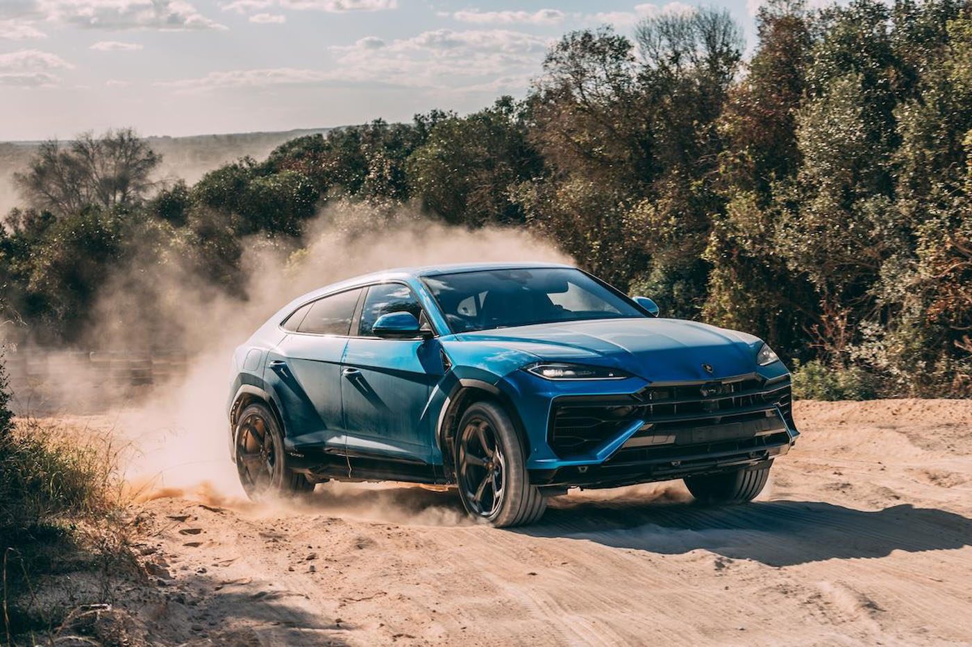 A dynamic debut unfolds as a blue SUV, akin to the hybrid Lamborghini Urus, powers through sandy terrain at Nardò, kicking up clouds of dust against a backdrop of trees and a moody sky.